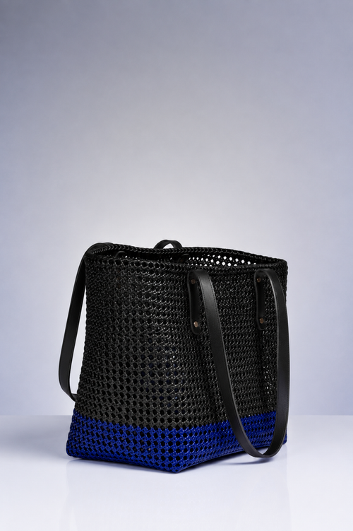 A black woven tote bag with a vibrant blue bottom section and black leather shoulder straps, photographed on a light gray studio background.