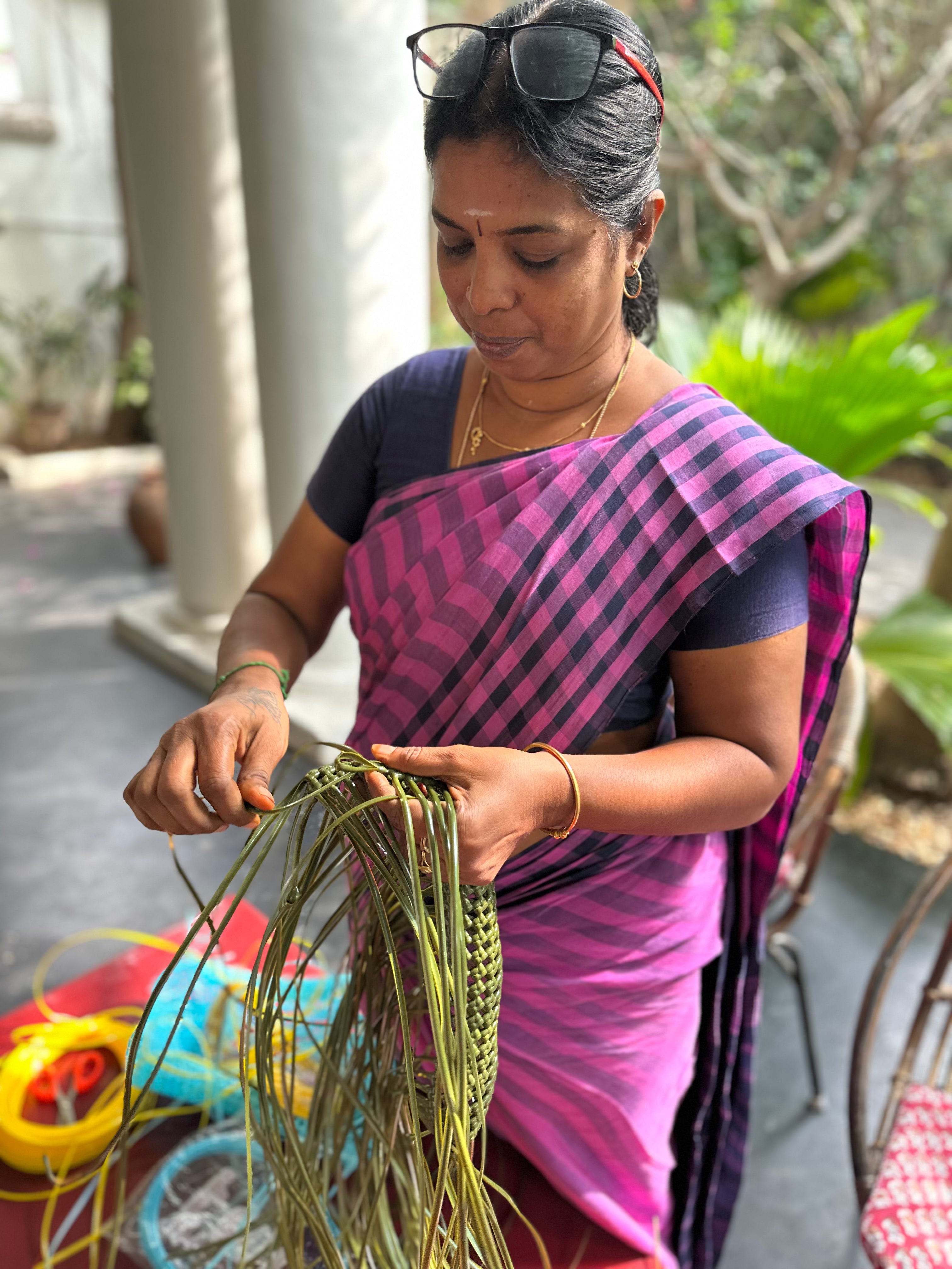 A woman wearing a pink and purple patterned sari is shown outdoors, crafting a woven green fiber bag with her hands. She has sunglasses on her head and gold jewelry on, with colorful crafting materials visible on the ground nearby.
