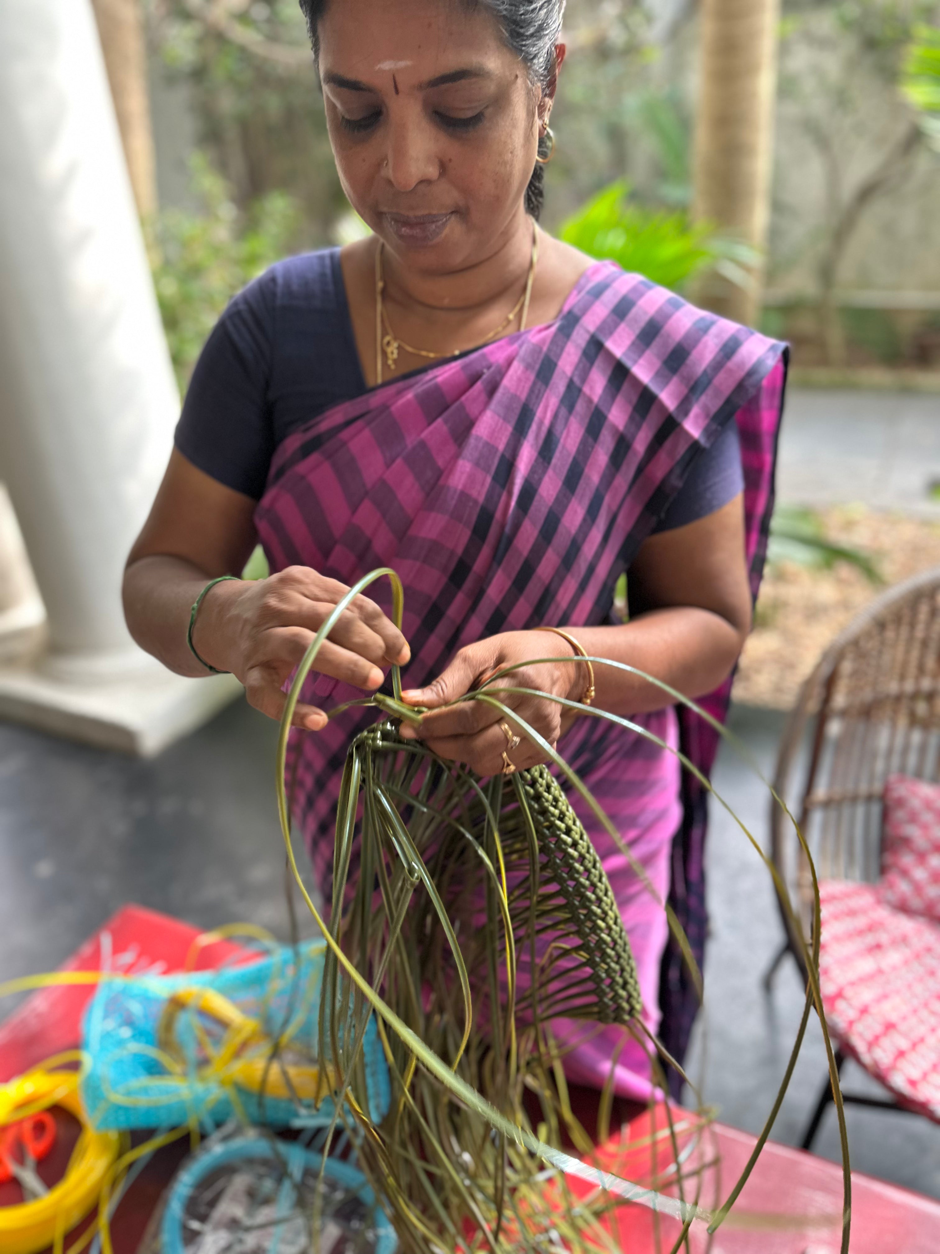 A woman wearing a purple and pink checkered sari is shown weaving a green  basket on a patio.