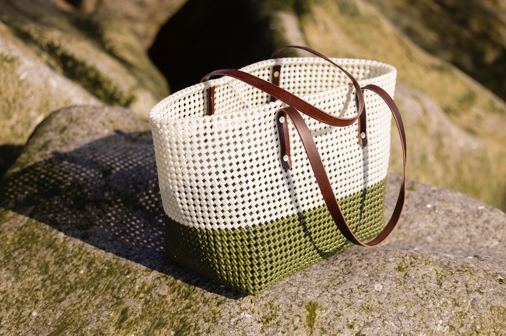 A woven basket bag with a white top half, olive green bottom half, and dark brown leather handles, resting on a mossy rock outdoors.