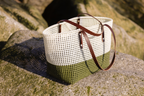 A woven basket bag with a white top half, olive green bottom half, and dark brown leather handles, resting on a mossy rock outdoors.