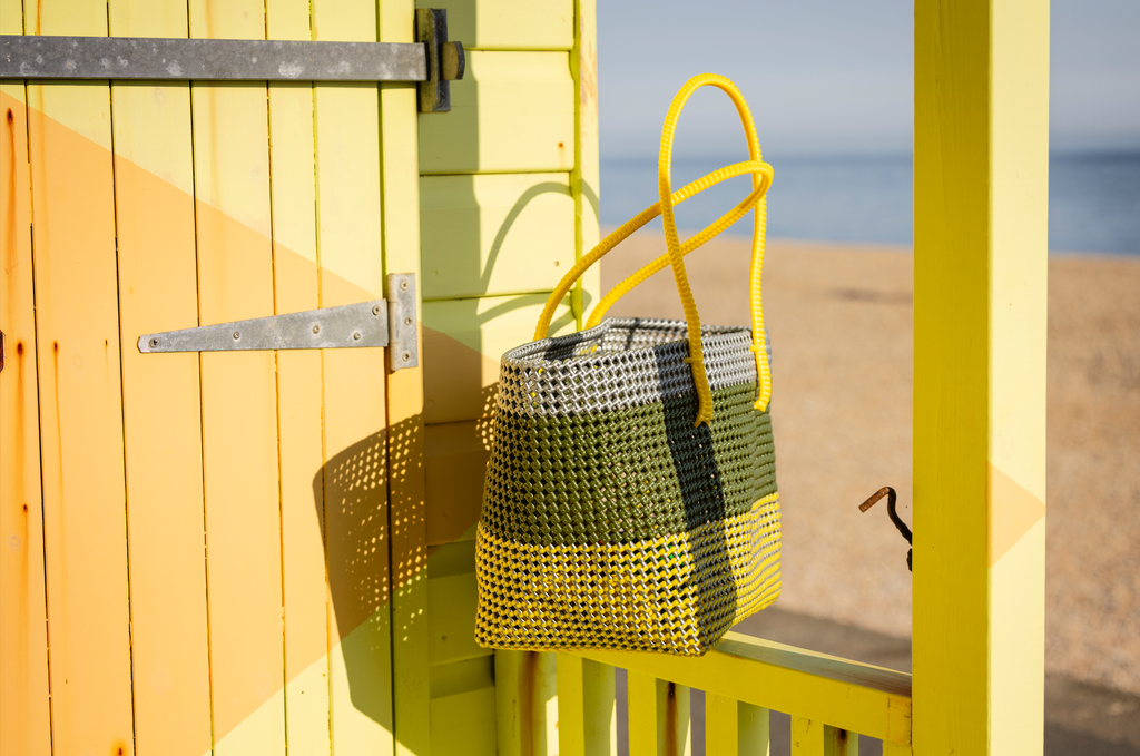 A woven tote bag with yellow, green, and black plaid stripes hangs on a yellow beach hut door, with a sandy beach and blue sea visible in the background.