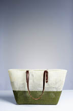 A woven tote bag with a two-tone design, featuring a cream upper section and an olive green lower section, with brown leather handles, set against a studio background.