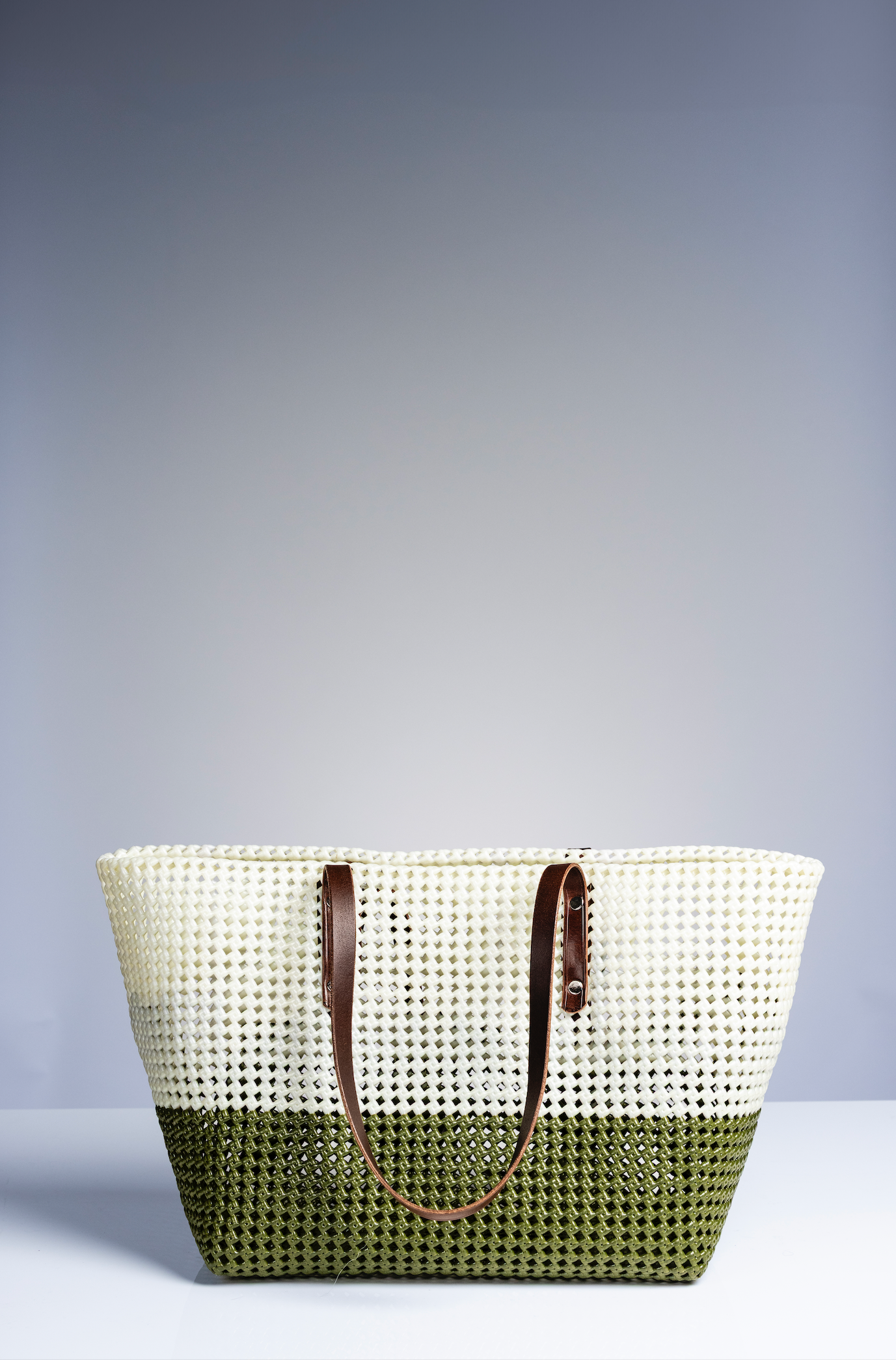 A woven tote bag with a two-tone design, featuring a cream upper section and an olive green lower section, with brown leather handles, set against a studio background.