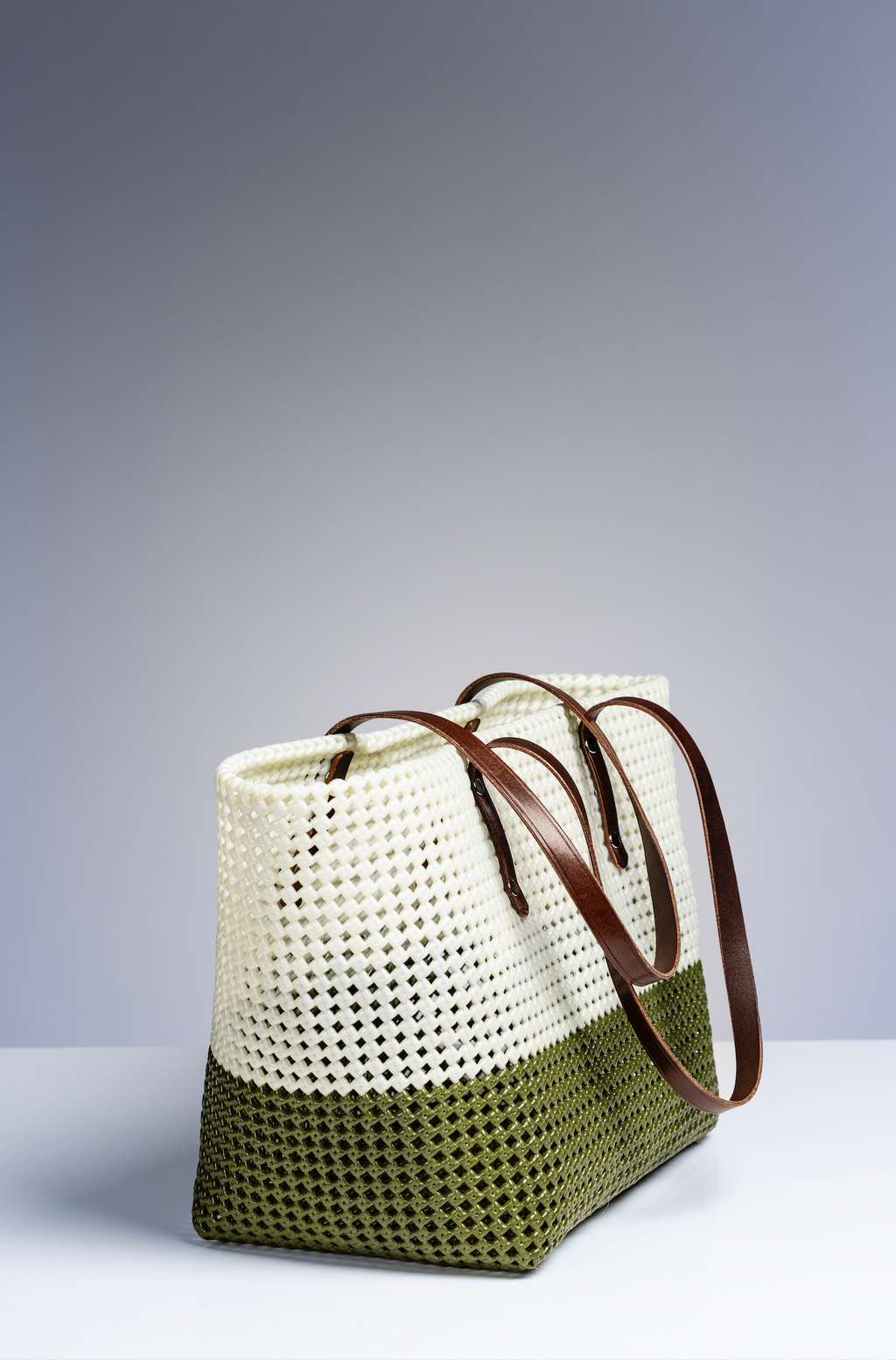 A woven tote bag with a two-tone design, featuring an off-white top section and an olive green bottom section, with brown leather shoulder straps, set against a neutral gray studio background.