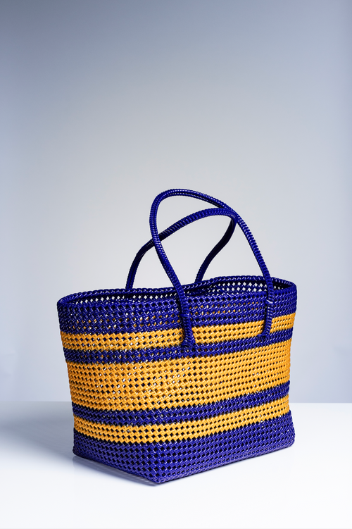 A woven basket bag with horizontal stripes in royal blue and golden yellow, featuring two top handles, set against a neutral gray studio background.