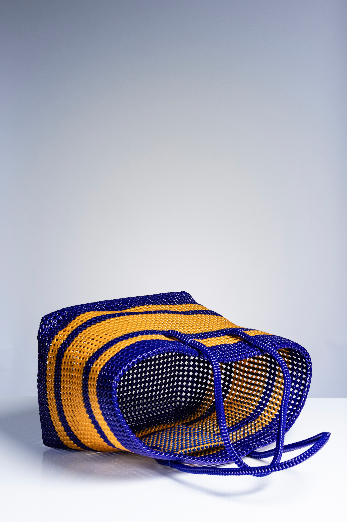 A woven basket bag with bold horizontal stripes in royal blue and mustard yellow, resting on a white surface against a light gray gradient background.