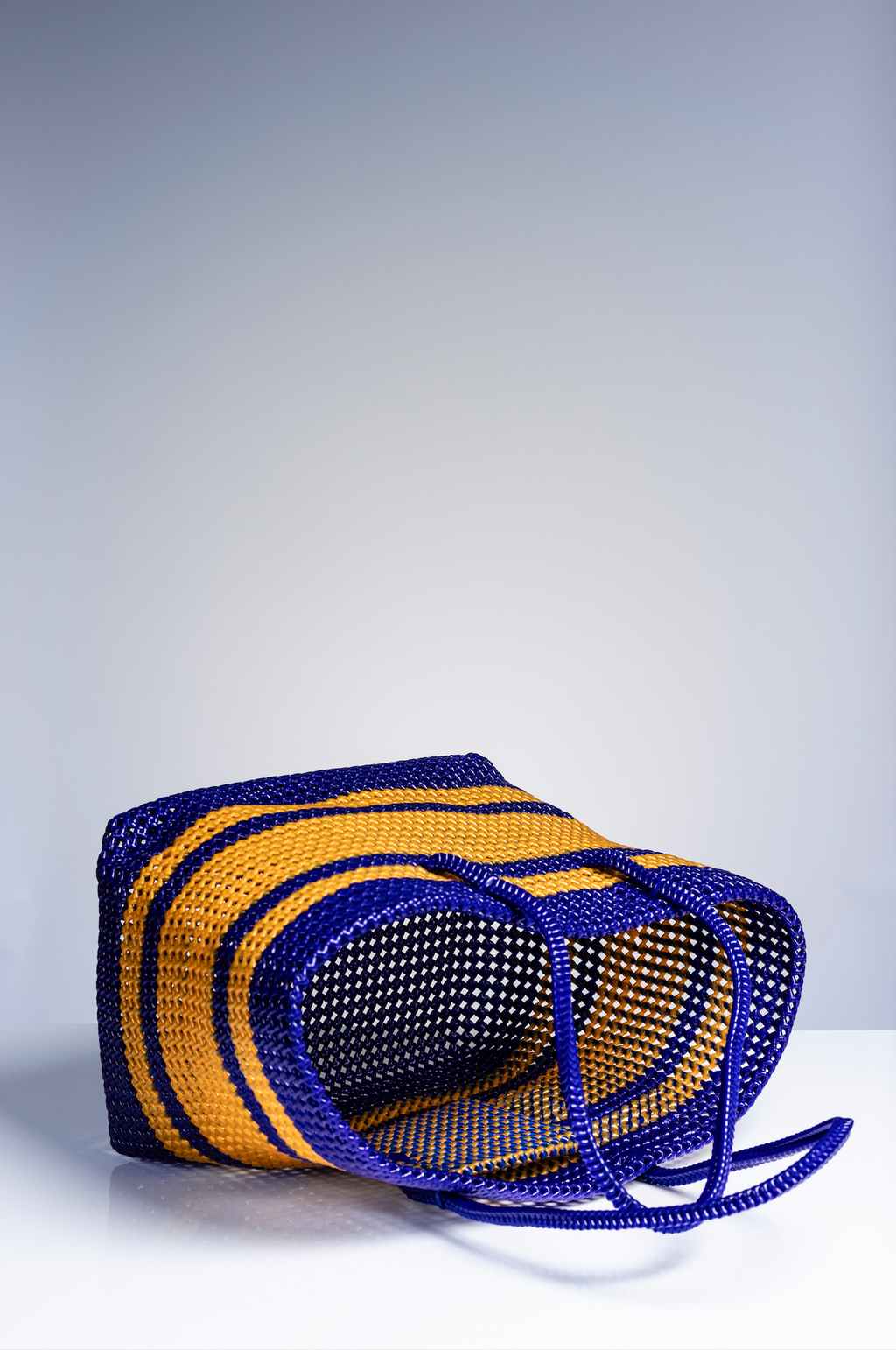 A woven basket bag with bold horizontal stripes in royal blue and mustard yellow, resting on a white surface against a light gray gradient background.