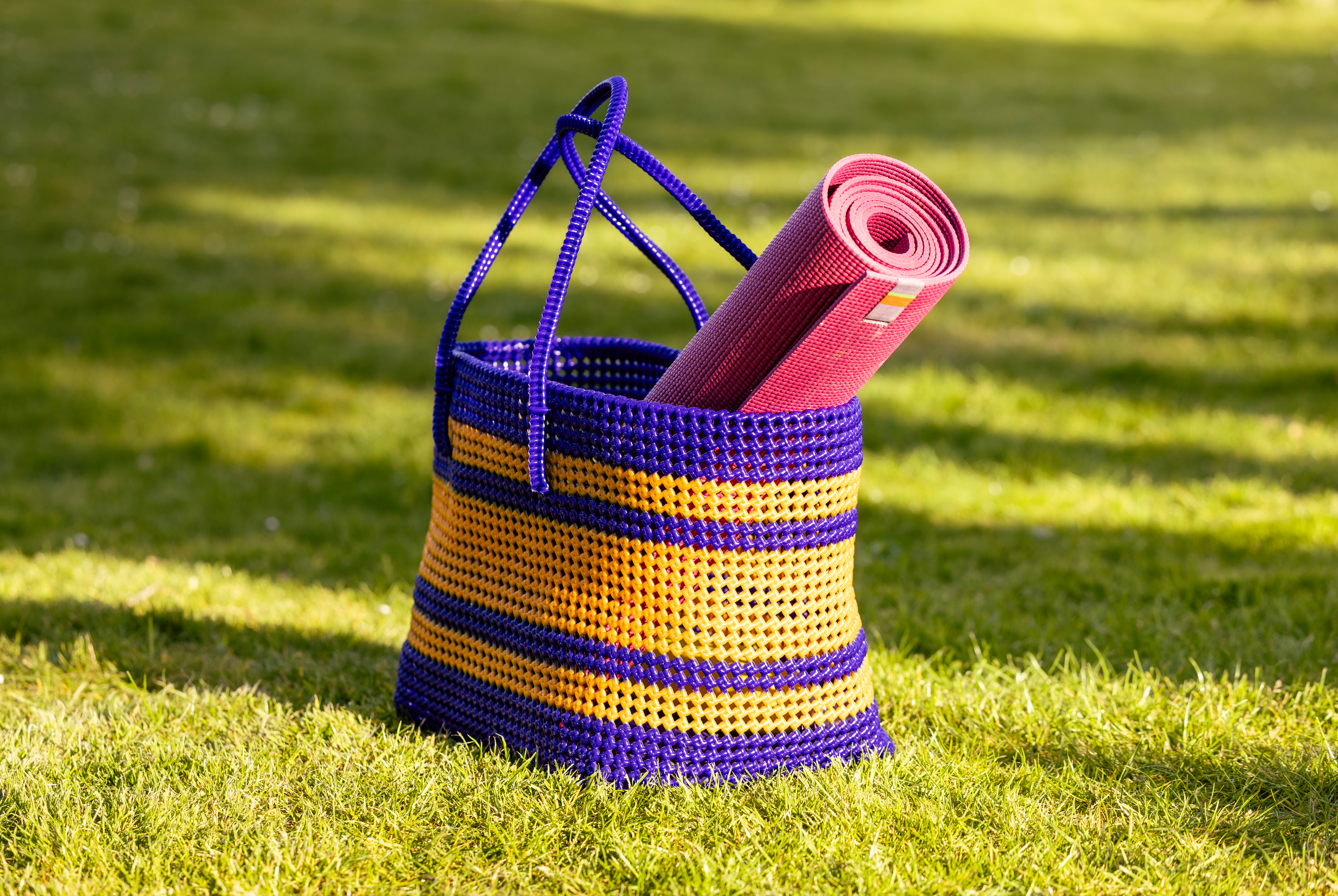 A blue and yellow woven basket bag with blue handles sits on a green grassy lawn, with a rolled pink yoga mat tucked inside.