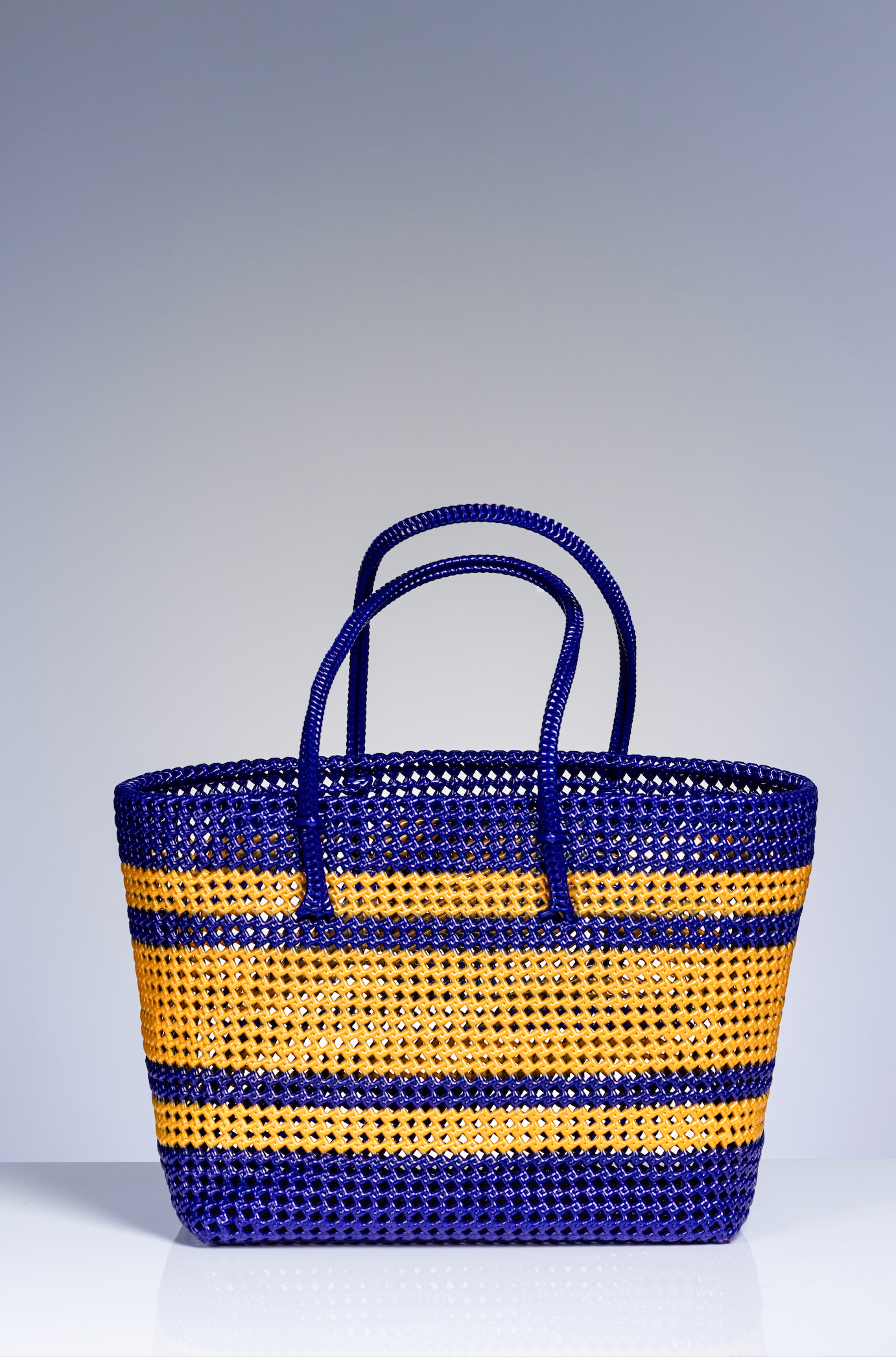 A woven basket bag with horizontal stripes in royal blue and golden yellow, featuring two top handles, set against a neutral gray studio background.