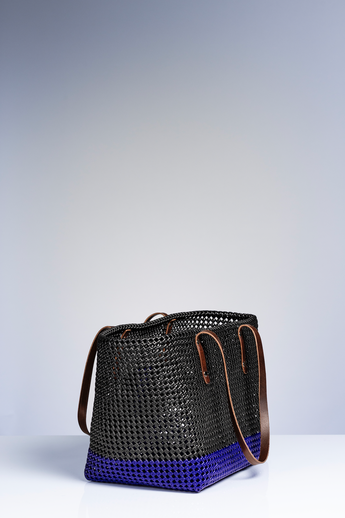 A woven basket bag with a black body and a vibrant blue bottom section, featuring brown leather shoulder straps, photographed on a light gray studio background.