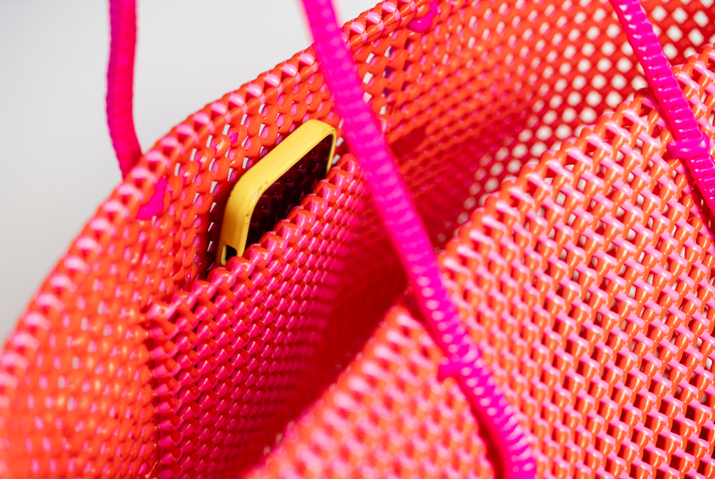 A close-up of a bright neon pink woven tote bag with a yellow phone partially tucked into one of its side pockets.