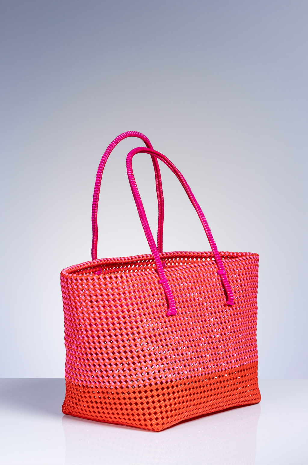 A woven tote bag with a bright orange base and a vibrant pink gradient top and handles, set against a light gray studio background.