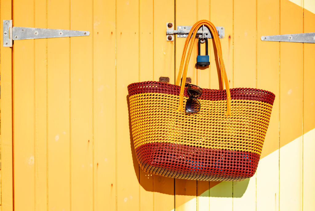 A woven tote bag with yellow and reddish-brown horizontal stripes hangs from a metal hook on a yellow wooden door. Sunglasses are tucked into one of the handles, and a blue padlock is hanging from the hook.