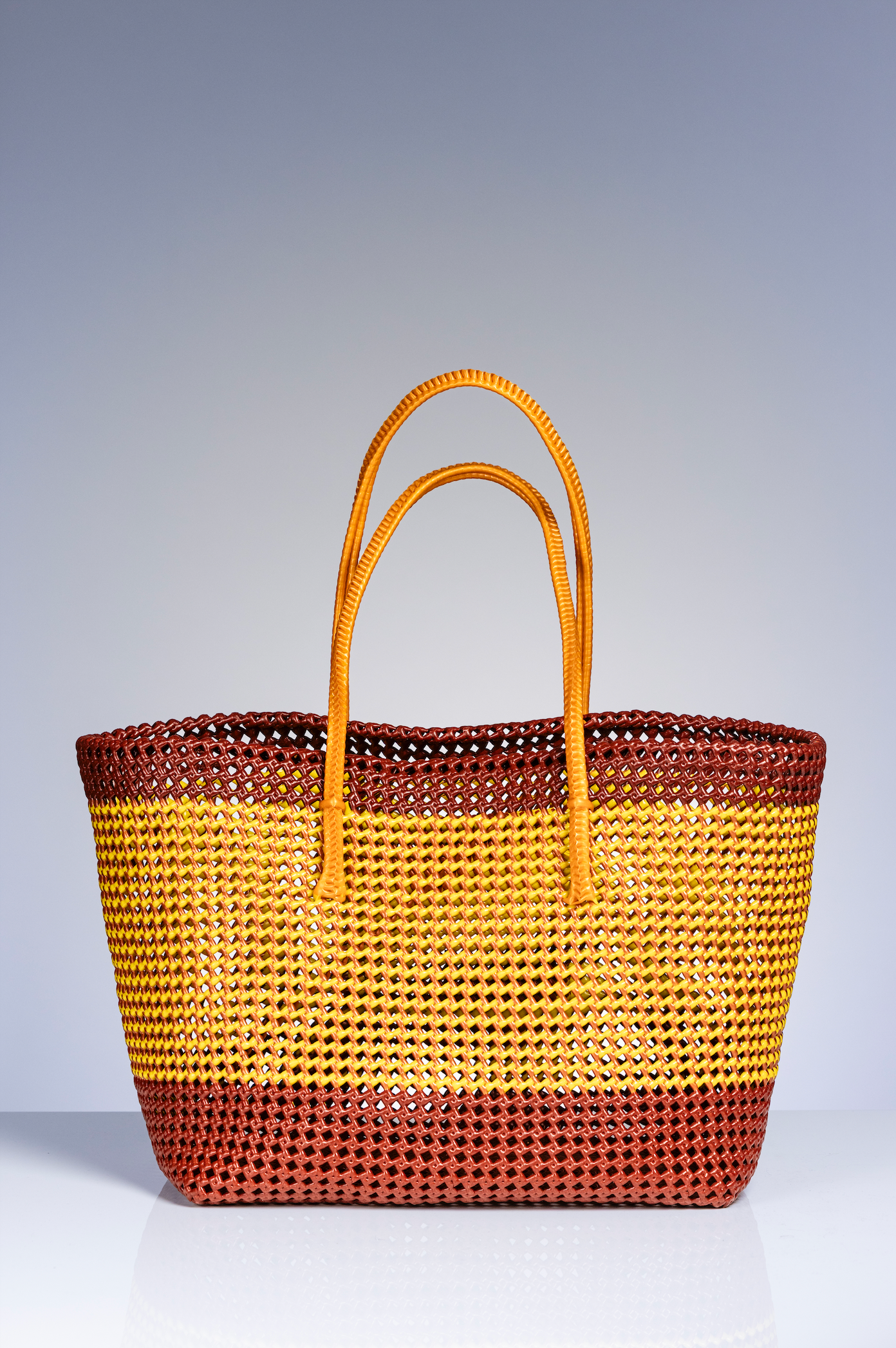 A woven tote bag with horizontal stripes in yellow, brown, and dark reddish-brown, featuring two tan shoulder straps, set against a gradient gray studio background.