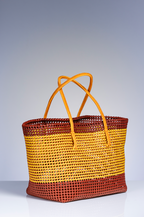 A woven straw tote bag with horizontal stripes in brown and golden yellow, featuring two tan leather shoulder straps, set against a neutral gray studio background.