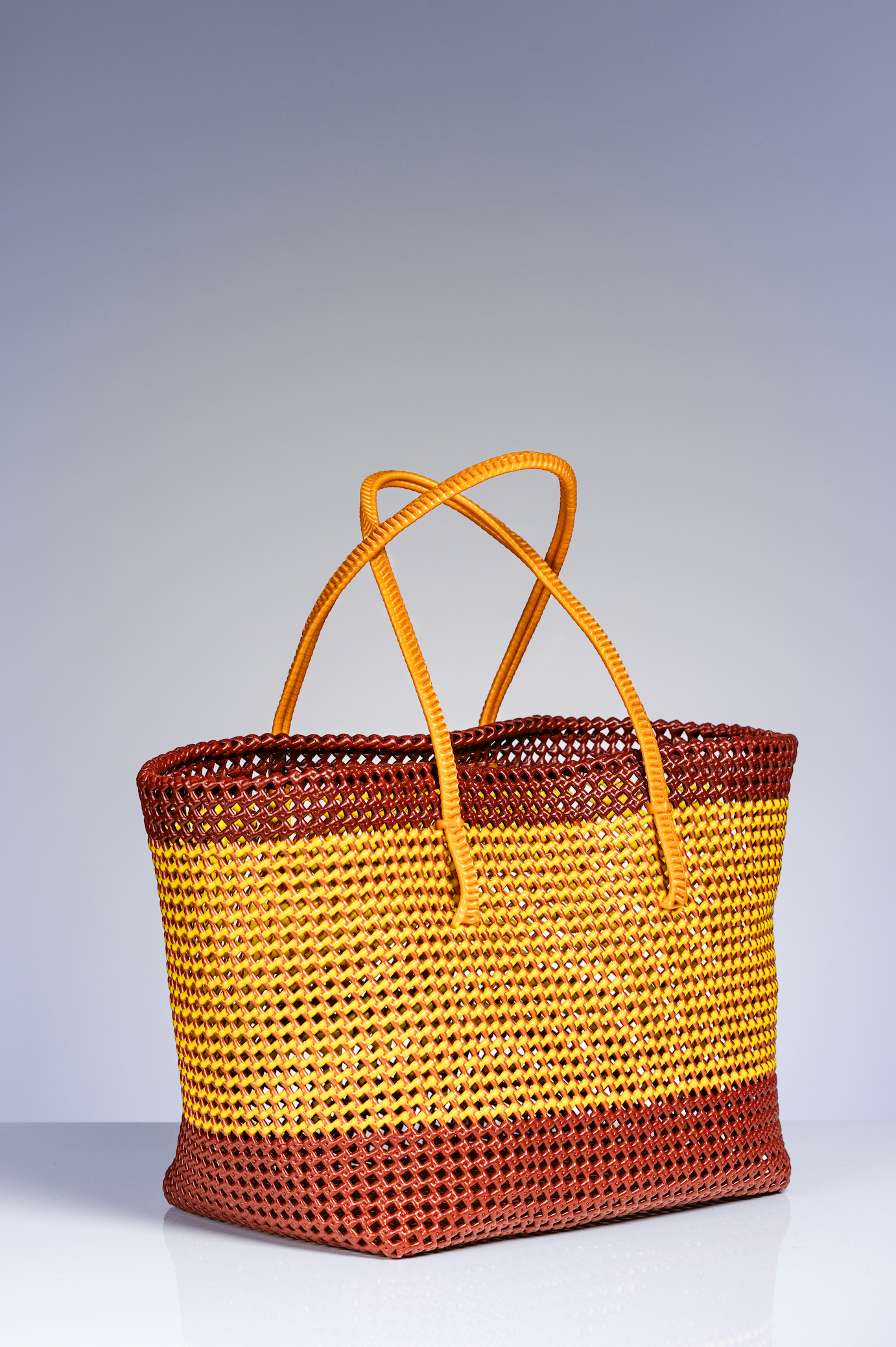A woven straw tote bag with horizontal stripes in brown and golden yellow, featuring two tan leather shoulder straps, set against a neutral gray studio background.