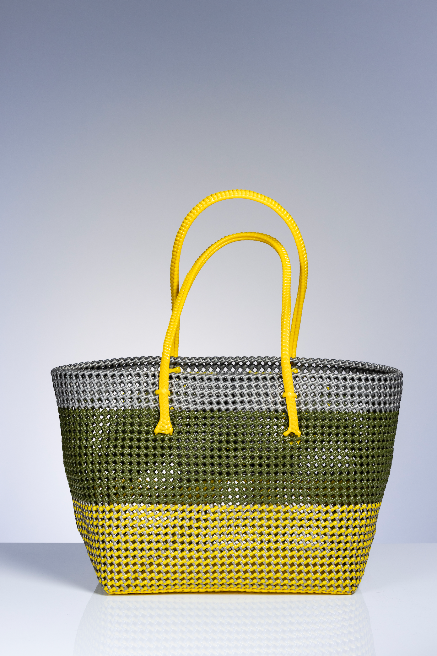 A woven straw tote bag with a three-tone color-blocked design in olive green, gray, and bright yellow, featuring matching yellow handles, set against a gradient gray studio background.