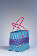 A woven tote bag with a light blue and purple striped body and bright pink handles, photographed on a neutral gray studio background.