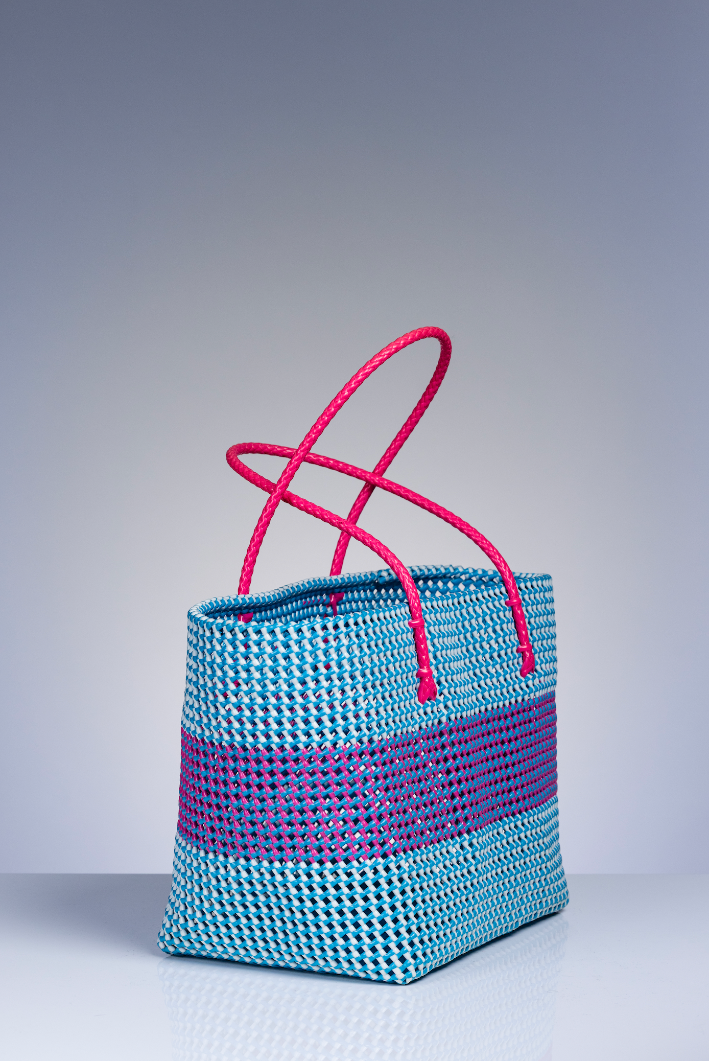 A woven tote bag with a light blue and purple striped body and bright pink handles, photographed on a neutral gray studio background.