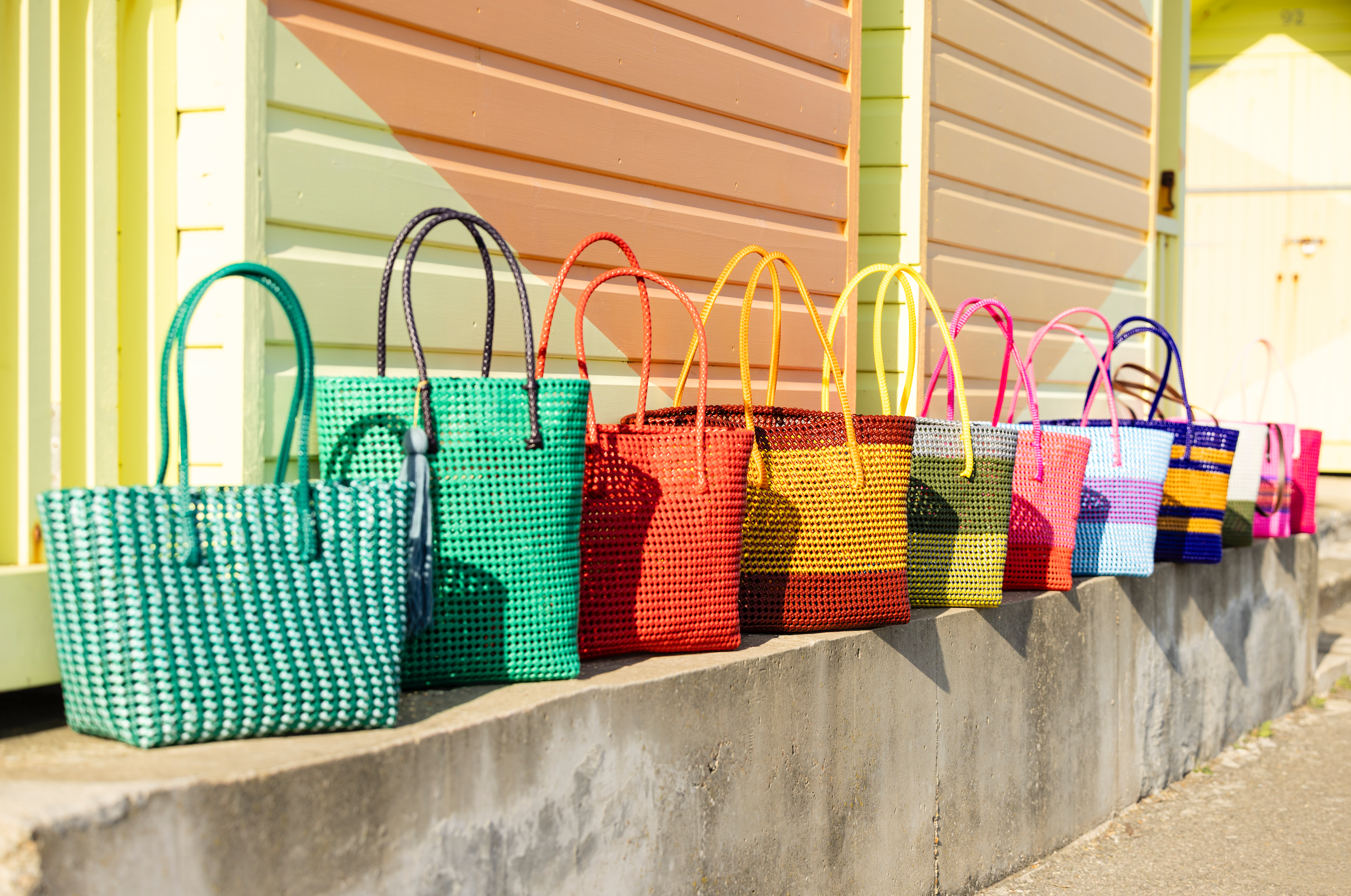 A row of colorful woven tote bags in different patterns and solid colors, lined up on a concrete ledge in front of a pastel-colored building.