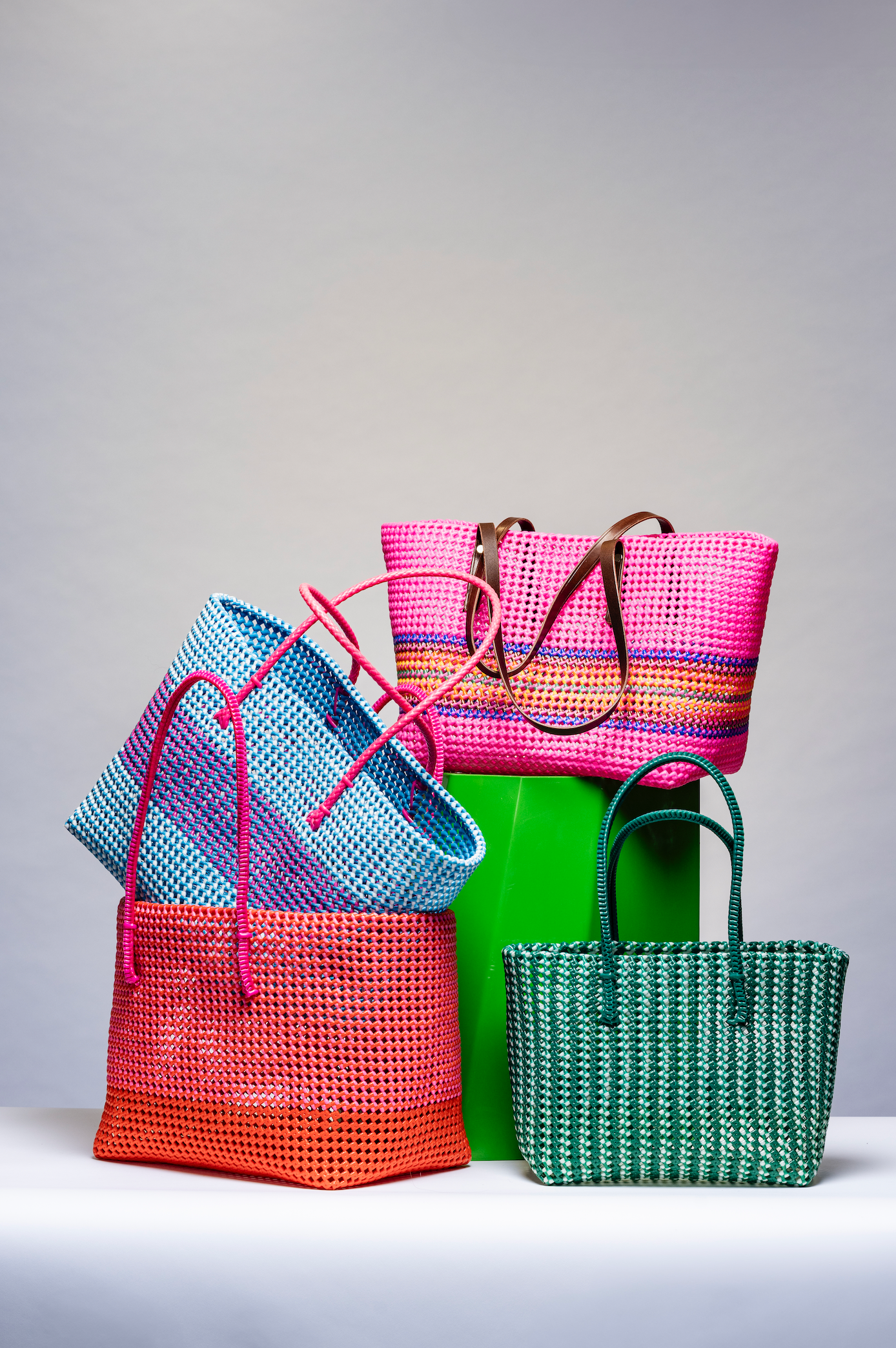 Four colorful woven tote bags arranged together on a light gray studio background. The bags include pink, blue, orange, green, and teal tones, with some stacked on top of each other.