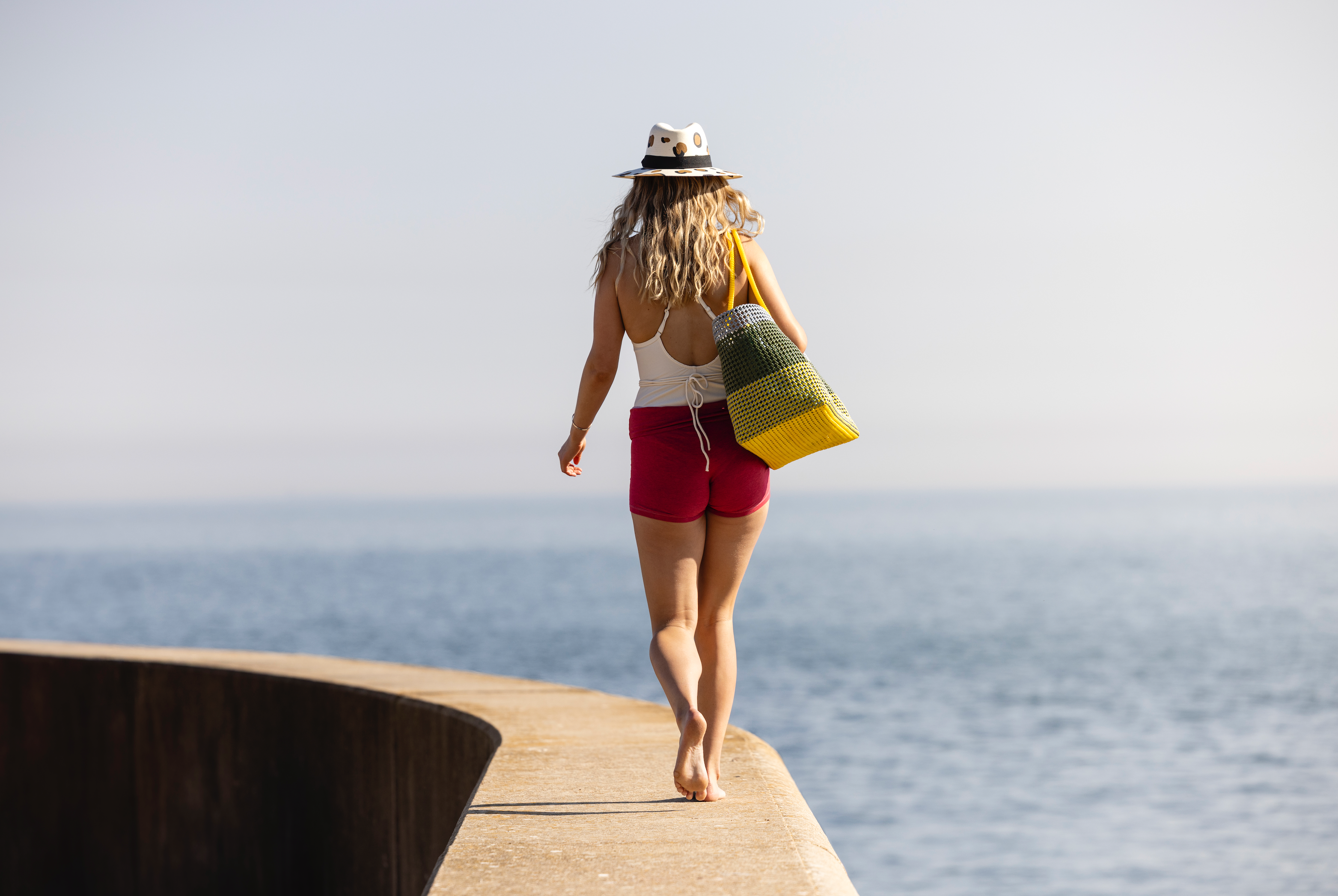 A woman seen from behind walking barefoot on a concrete pier with the ocean in the background, wearing a white hat, white tank top, red shorts, and carrying a yellow and green woven tote bag.